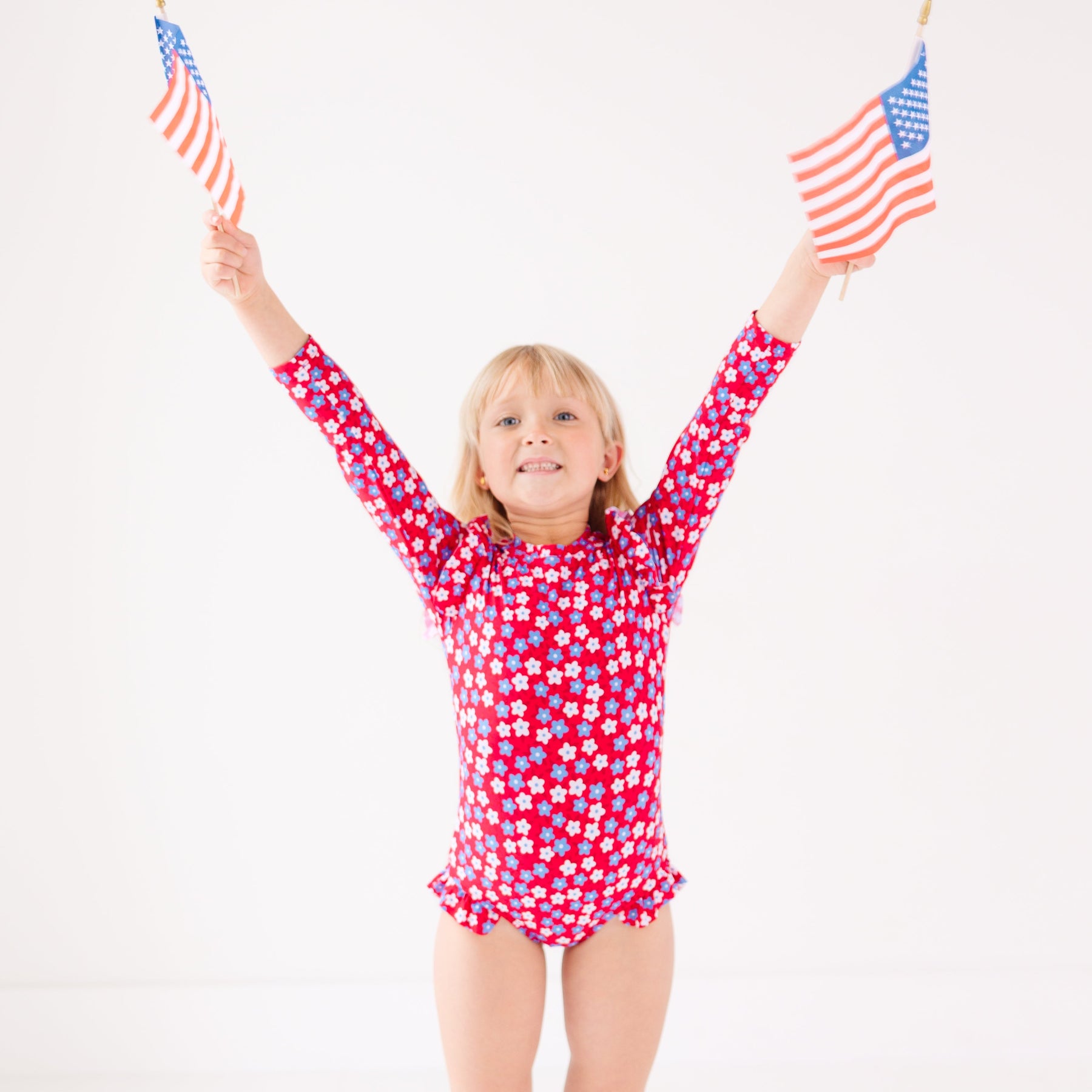 Young girl wearing a red floral swimsuit holding up two small American flags on a white background