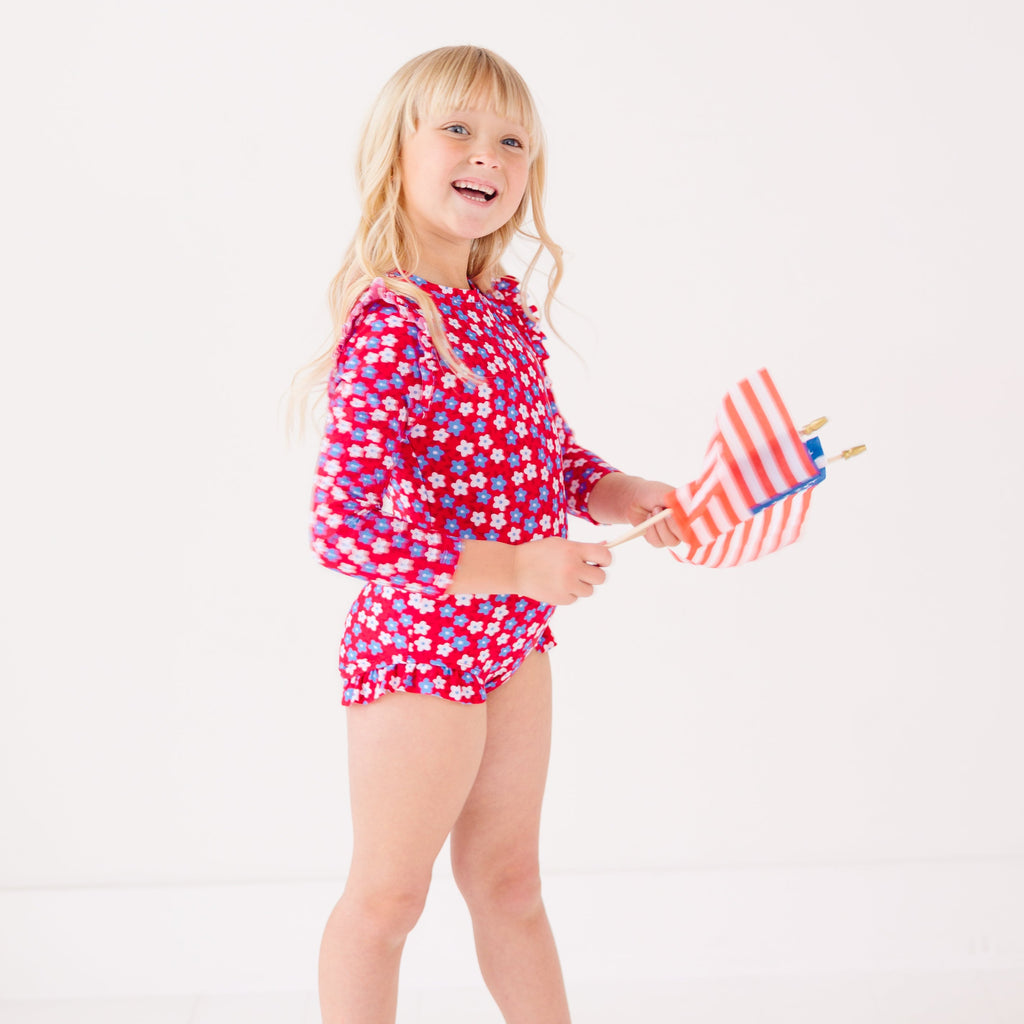 Young girl wearing a red floral swimsuit holding small American flags on a white background