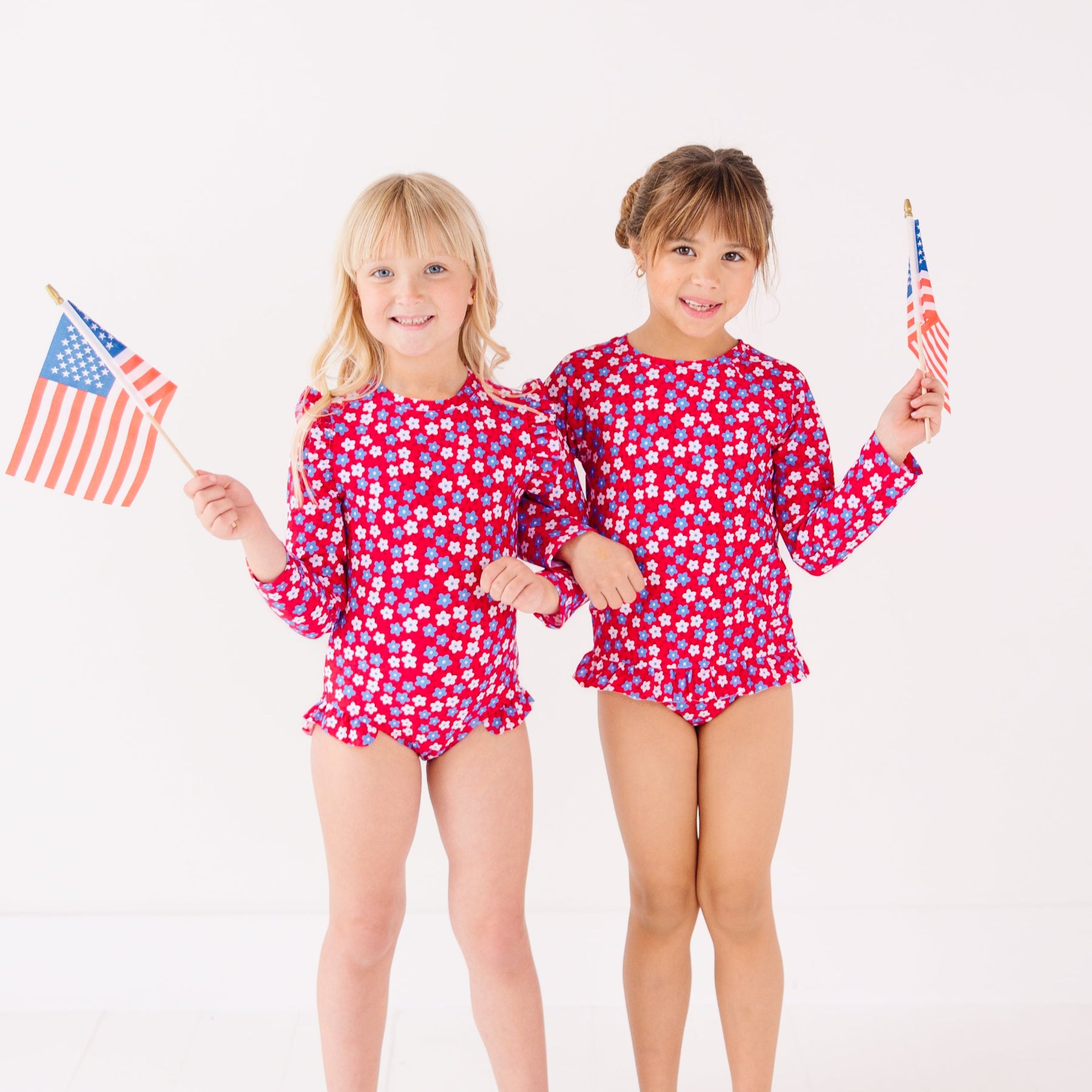 Two children in matching red and white swimsuits holding American flags on a white background