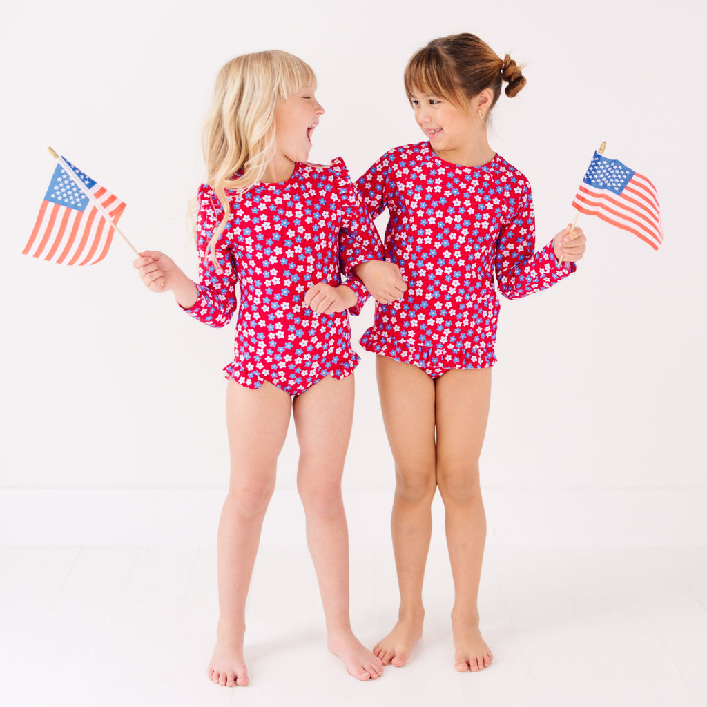 Two children wearing matching red and white floral patterned swimsuits, holding American flags, on a white background