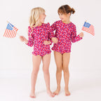 Two children wearing matching red and white floral patterned swimsuits, holding American flags, on a white background
