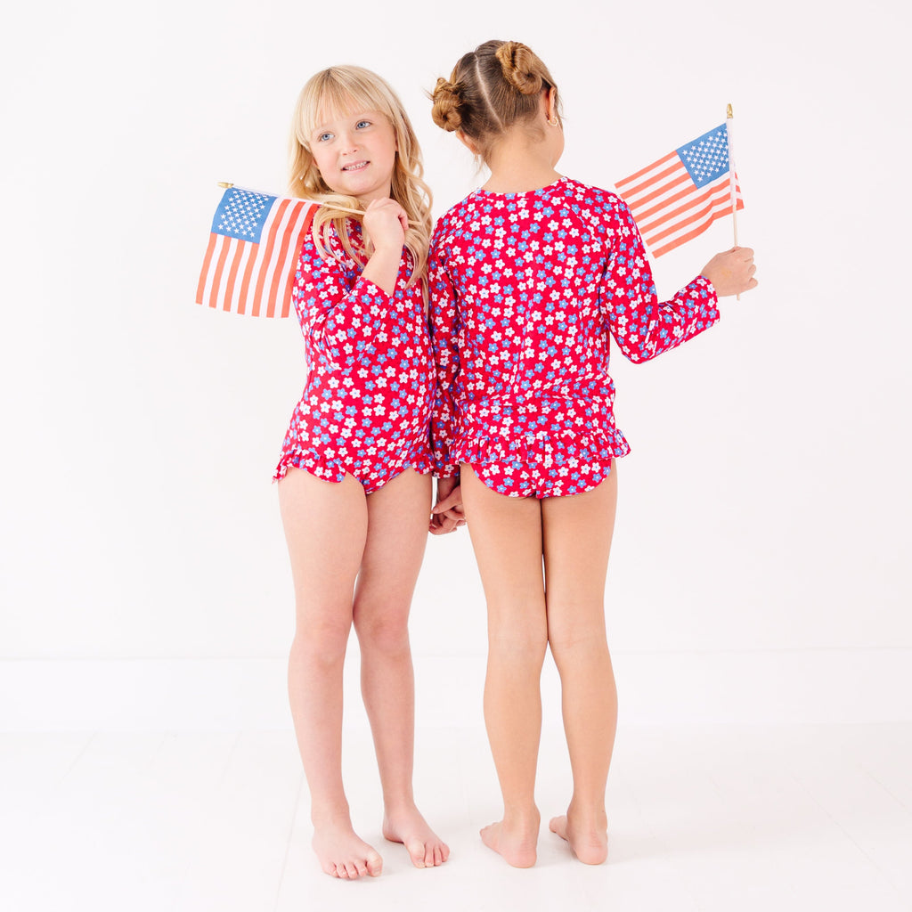 Two children wearing matching red and white floral patterned swimsuits, holding American flags, on a white background