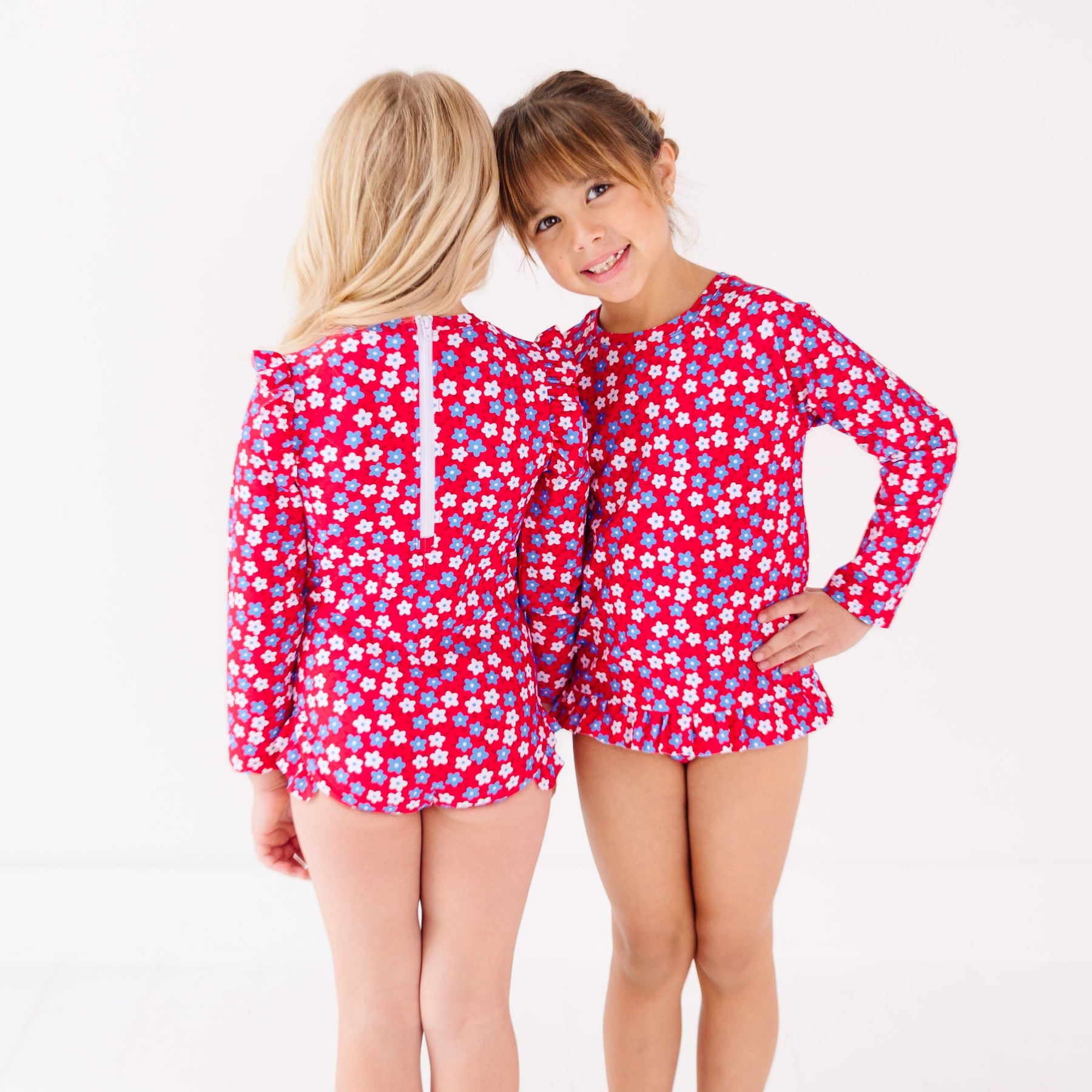 Two children wearing matching red and white floral patterned swimsuits on a white background