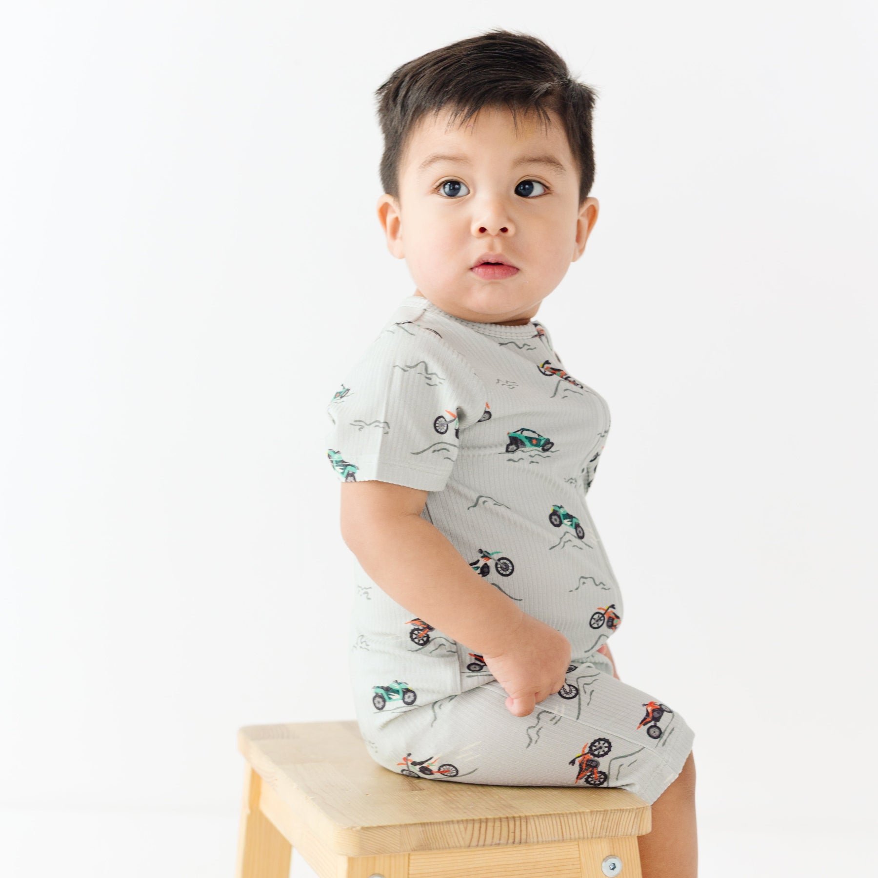 Baby wearing a light gray onesie with vehicle patterns sitting on a wooden stool against a white background