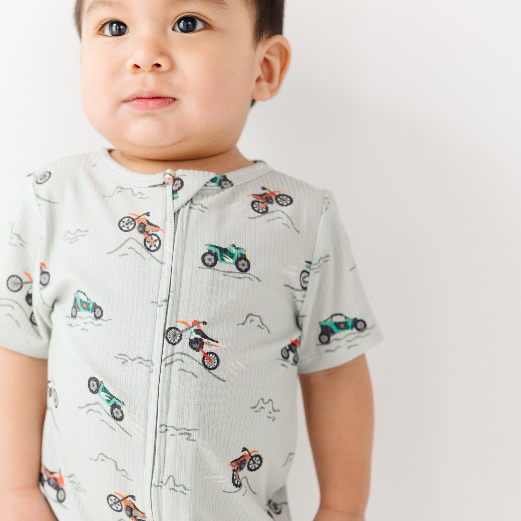 Baby wearing a light gray onesie with vehicle patterns on a white background