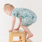 Child wearing a blue short romper with summer patterns climbing a stool