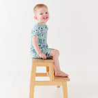 Child wearing a blue short romper with summer patterns sitting on a stool 