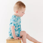 Child wearing a blue palm tree patterned onesie sitting on a wooden stool against a white background