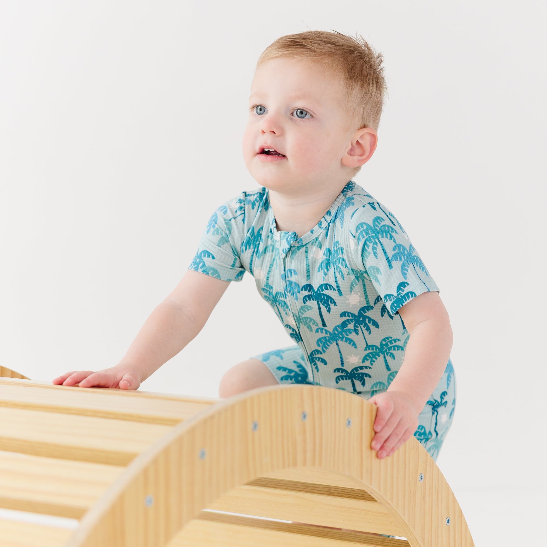 Child wearing a blue palm tree patterned onesie sitting on a wooden stool against a white background
