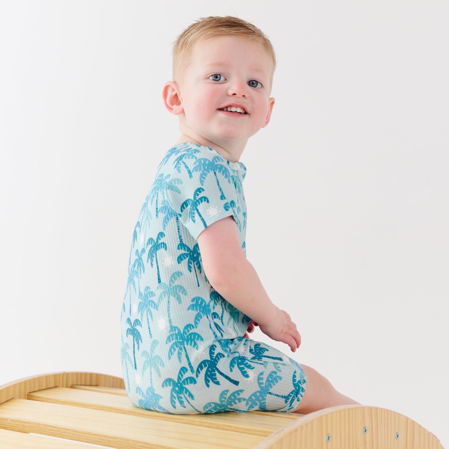 Child wearing a blue palm tree patterned onesie sitting on a wooden stool against a white background