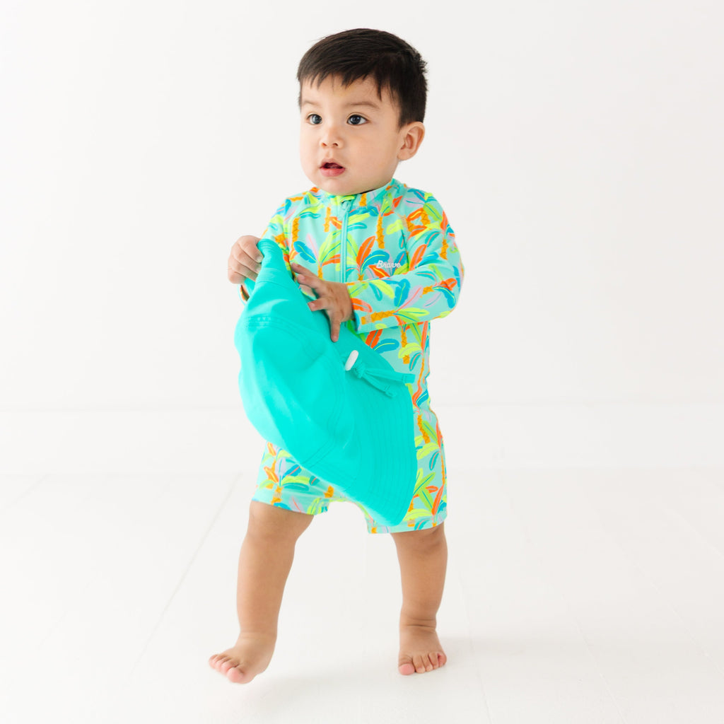 Baby wearing a colorful boy swimsuit with palm trees pattern, holding a turquoise bucket hat on a white background