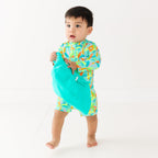 Baby wearing a colorful boy swimsuit with palm trees pattern, holding a turquoise bucket hat on a white background