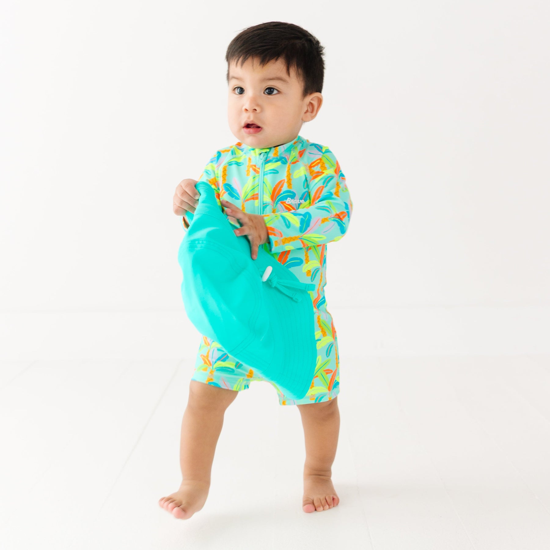Baby wearing a colorful boy swimsuit with palm trees pattern, holding a turquoise bucket hat on a white background