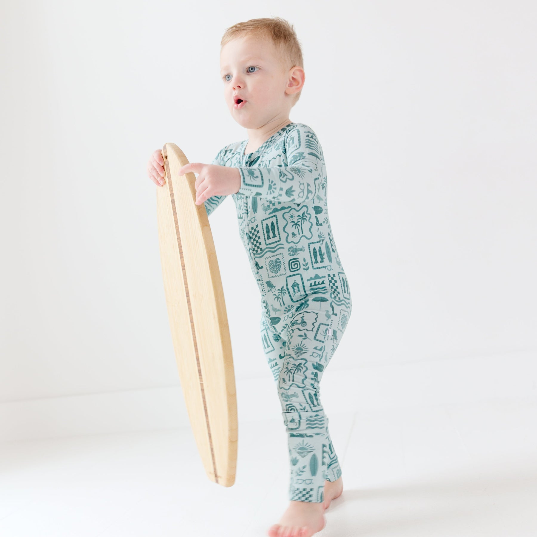 Child wearing a blue summer-patterned onesie holding a tiny surfboard on a white background
