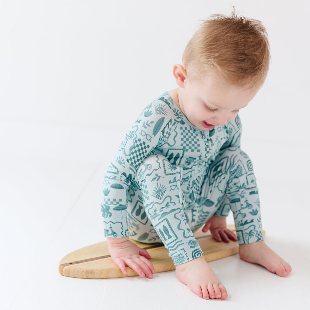 Child wearing a blue summer-patterned onesie sitting on a tiny surfboard on a white background