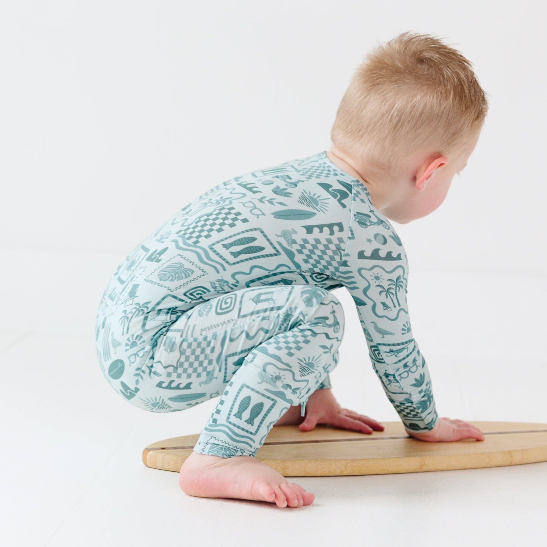 Child wearing a blue summer-patterned onesie sitting on a tiny surfboard on a white background