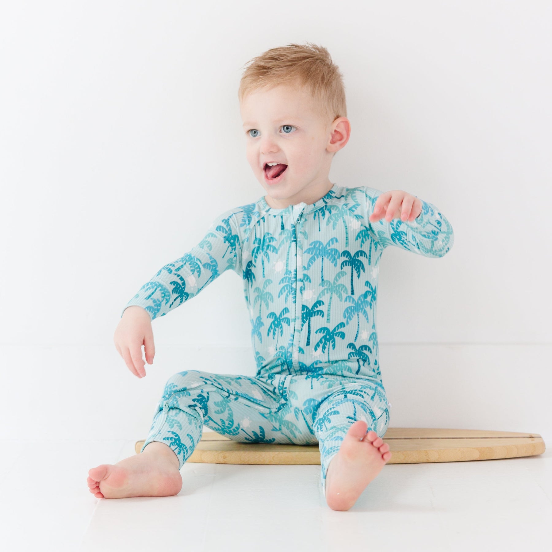 Child wearing a blue palm tree patterned onesie sitting on a surfboard against a white background