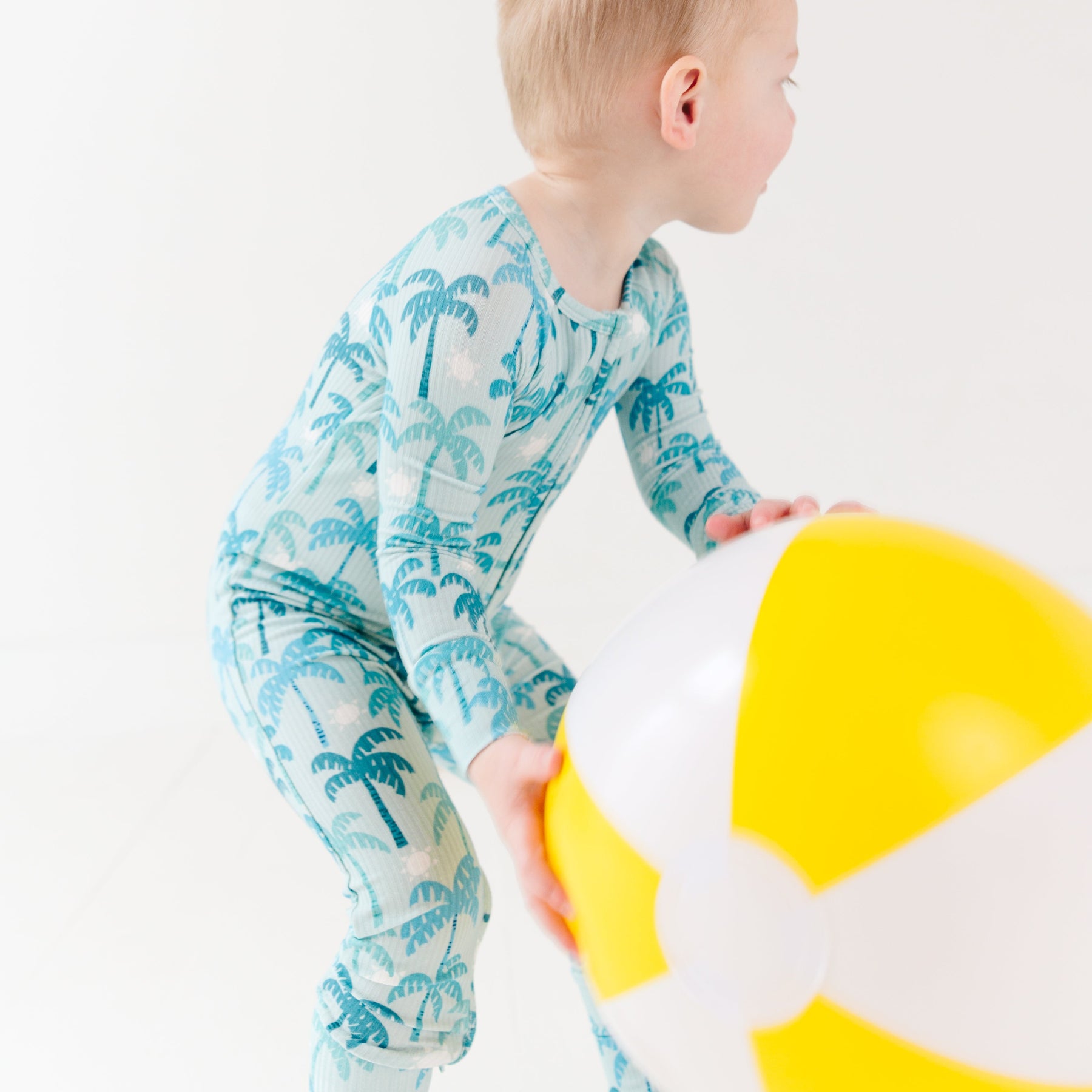 Child wearing a blue palm tree patterned onesie holding a beach ball on a white background