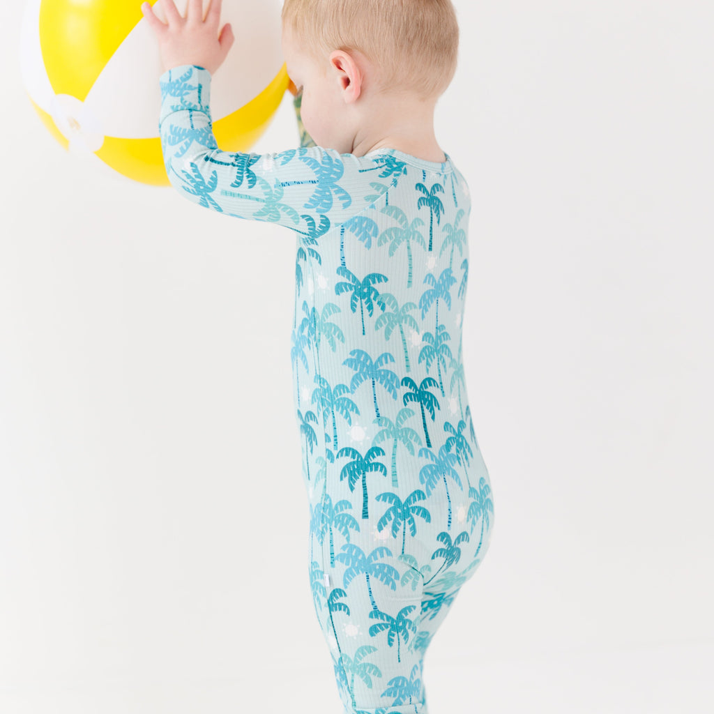 Child wearing a blue palm tree patterned onesie holding a beach ball on a white background