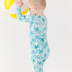 Child wearing a blue palm tree patterned onesie holding a beach ball on a white background