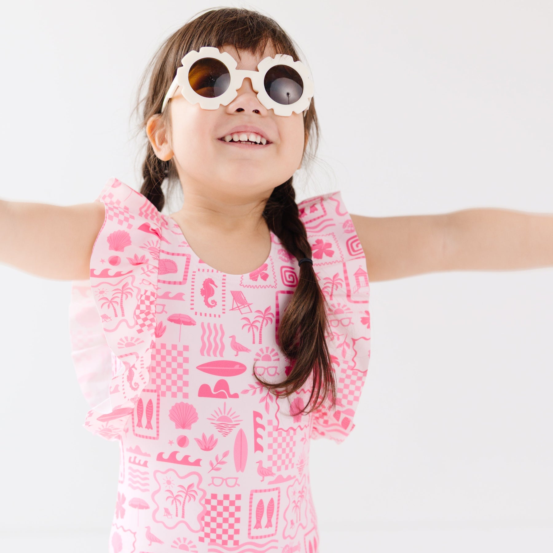 Child wearing a pink and white patterned ruffled one-piece swimsuit and flower sunglasses on a white background