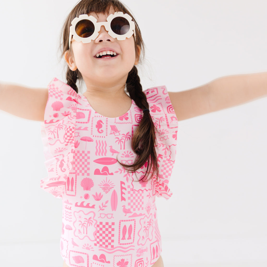 Child wearing a pink and white patterned ruffled one-piece swimsuit and flower sunglasses on a white background