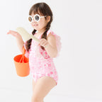 Child wearing a pink and white patterned ruffled one-piece swimsuit and flower sunglasses holding a sand bucket and shovel on a white background