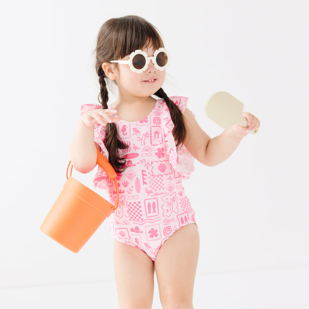 Child wearing a pink and white patterned ruffled one-piece swimsuit and flower sunglasses holding a sand bucket and shovel on a white background
