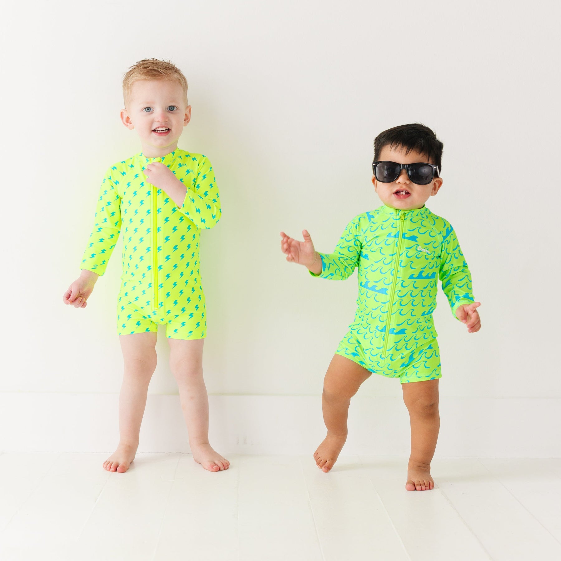 Two children wearing matching green boy swimsuits with waves and lightning bolt patterns on a white background.
