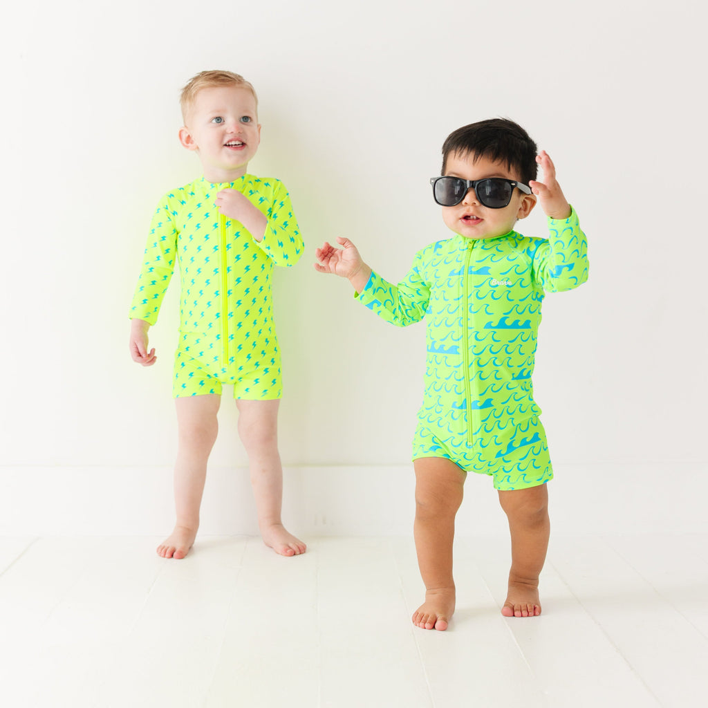 Two children wearing matching green boy swimsuits with waves and lightning bolt patterns on a white background.