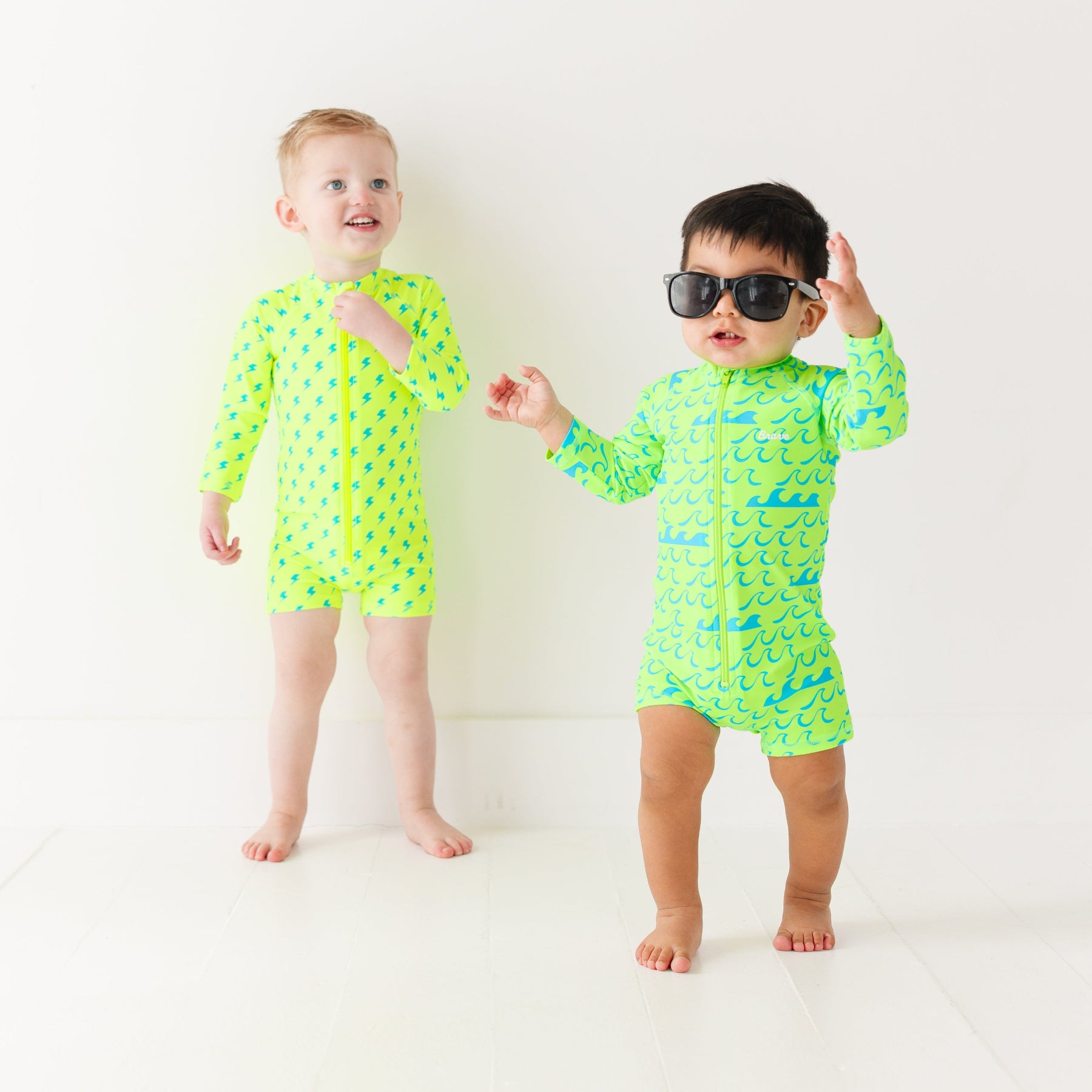 Two children wearing matching green boy swimsuits with waves and lightning bolt patterns on a white background.