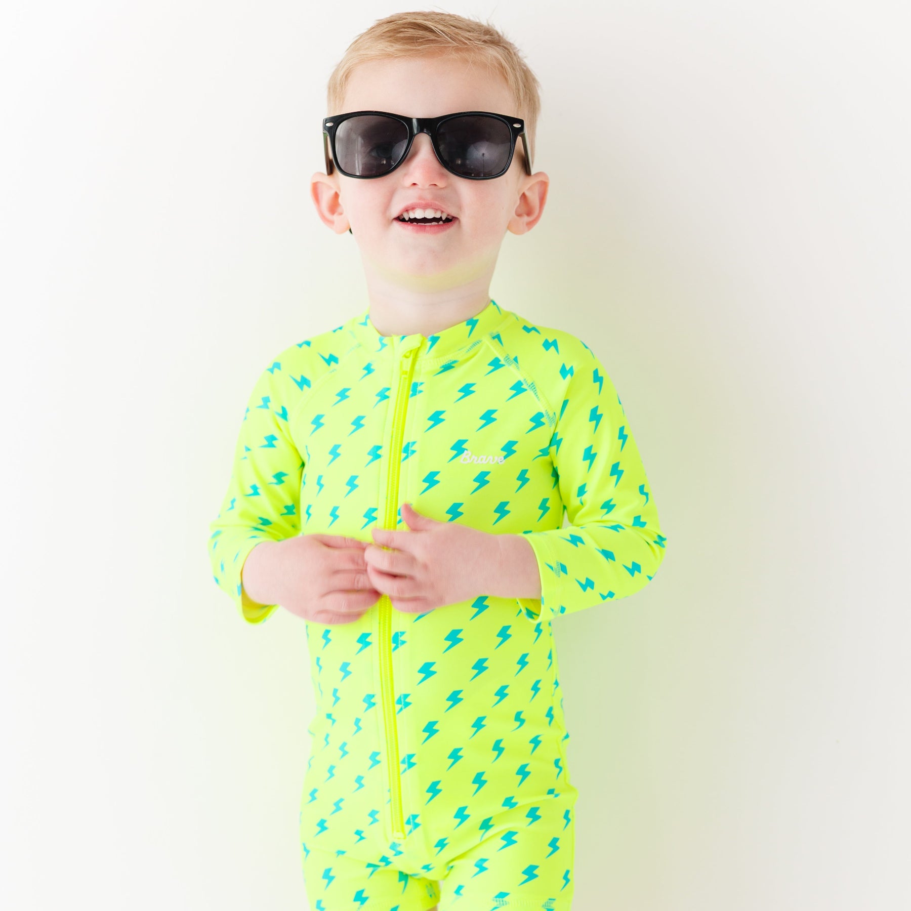Child wearing a bright yellow boy swimsuit with a lightning bolt pattern and sunglasses on a white background