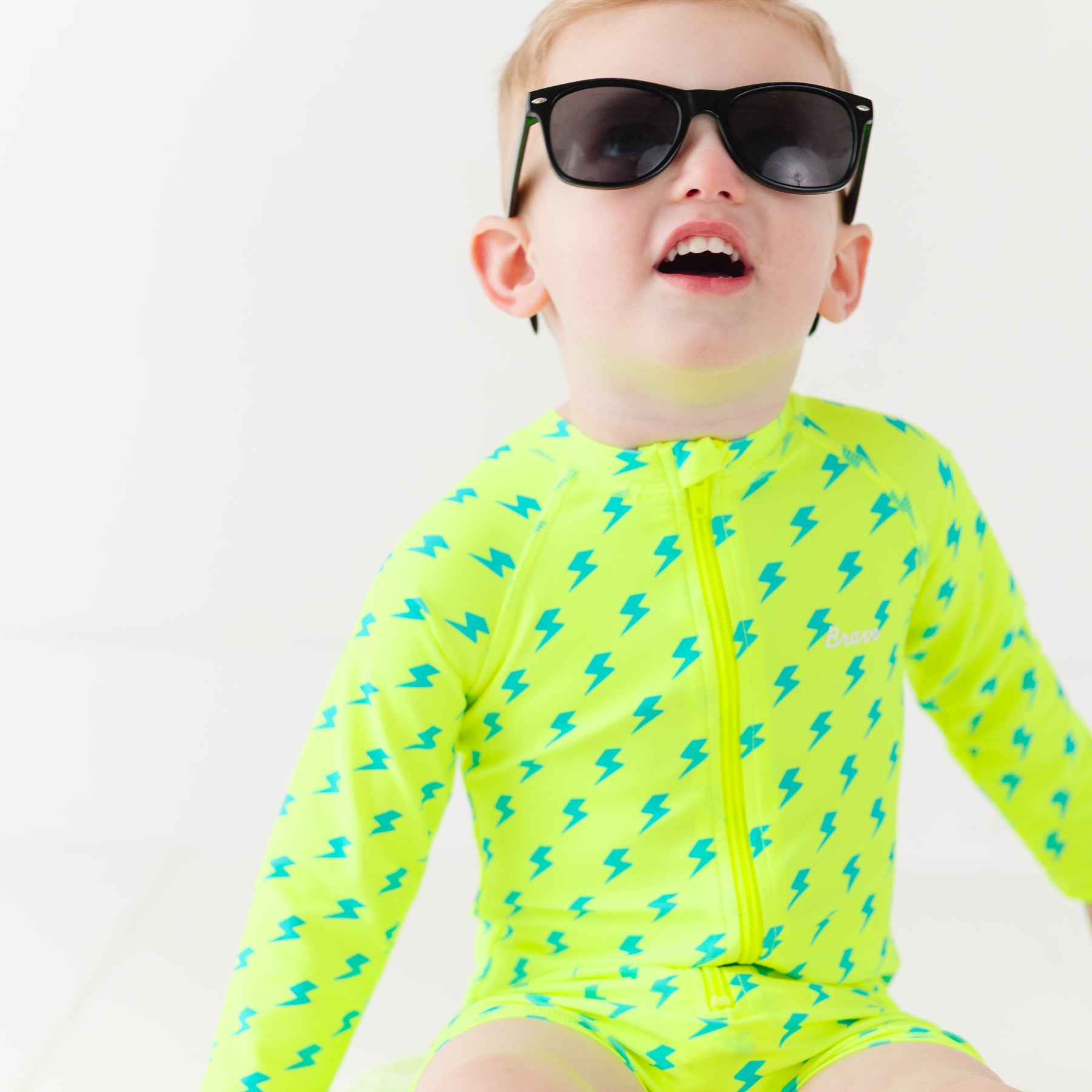 Child wearing a bright yellow boy swimsuit with a lightning bolt pattern and sunglasses on a white background