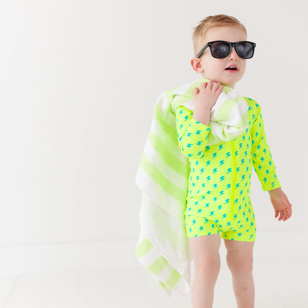 Child wearing a bright yellow boy swimsuit with a lightning bolt pattern and sunglasses on a white background, holding a green and white-striped towel