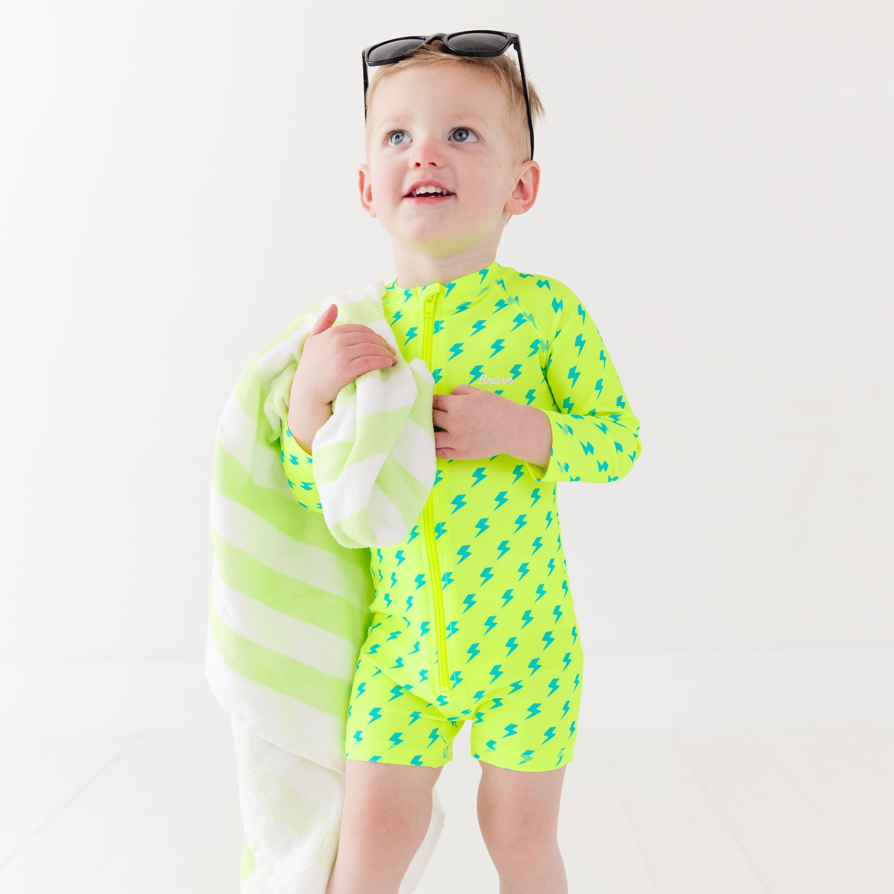 Child wearing a bright yellow boy swimsuit with a lightning bolt pattern and sunglasses on a white background, holding a green and white-striped towel