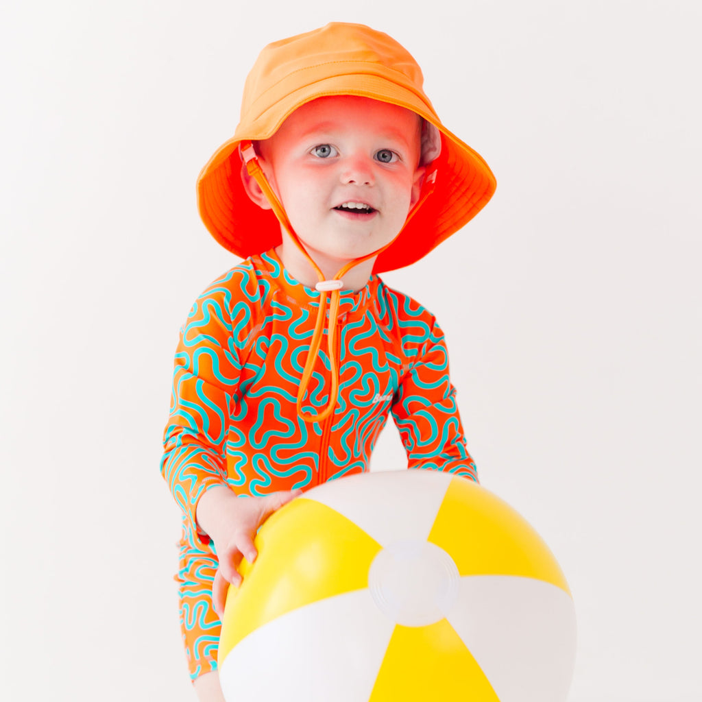 Child wearing a colorful swimsuit with a squiggly pattern and an orange bucket hat on a white background, holding a white and yellow-striped beach ball