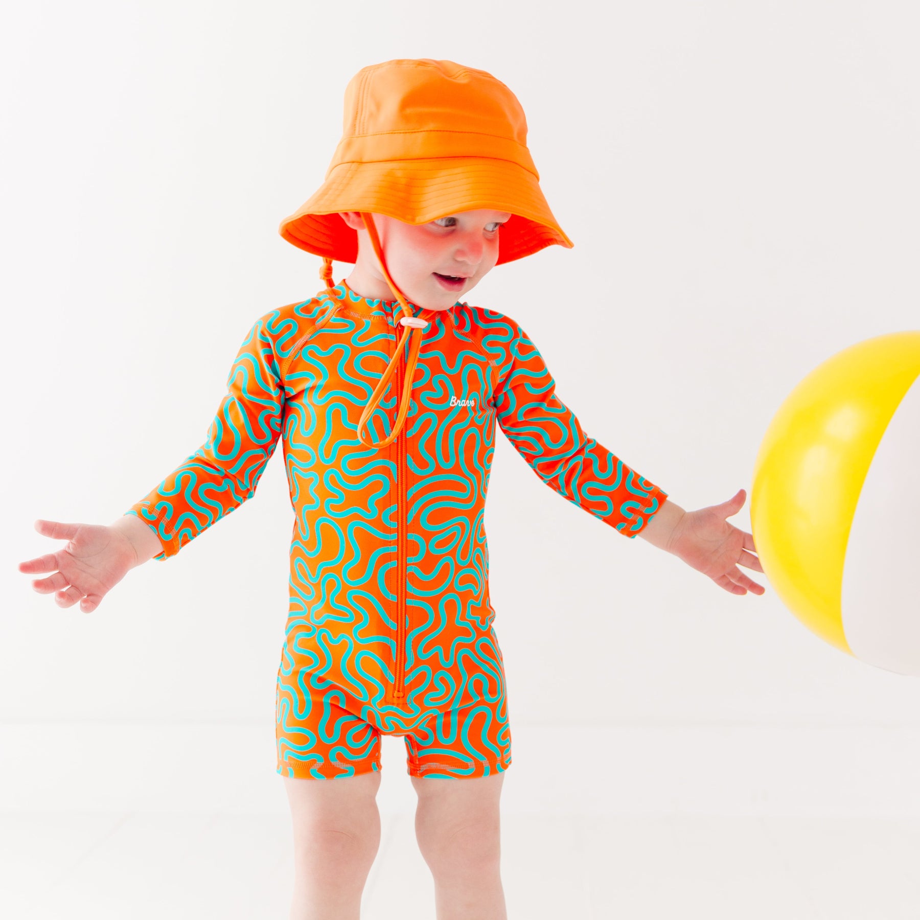 Child wearing a colorful swimsuit with a squiggly pattern and an orange bucket hat on a white background, holding a white and yellow-striped beach ball
