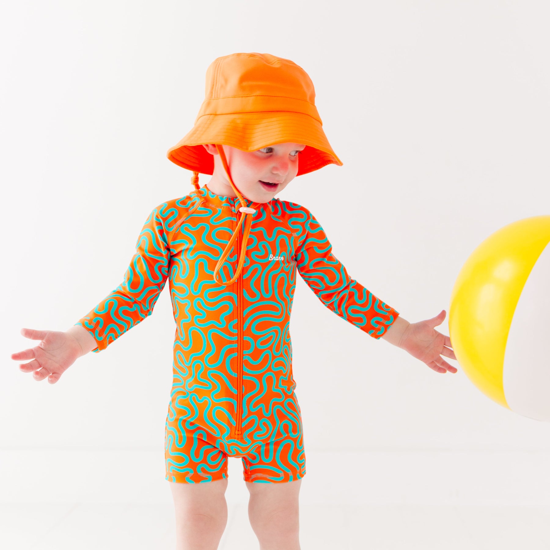 Child wearing a colorful swimsuit with a squiggly pattern and an orange bucket hat on a white background, holding a white and yellow-striped beach ball