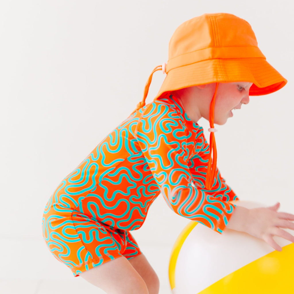 Child wearing a colorful swimsuit with a squiggly pattern and an orange bucket hat on a white background, holding a white and yellow-striped beach ball