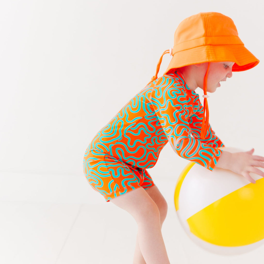 Child wearing a colorful swimsuit with a squiggly pattern and an orange bucket hat on a white background, holding a white and yellow-striped beach ball