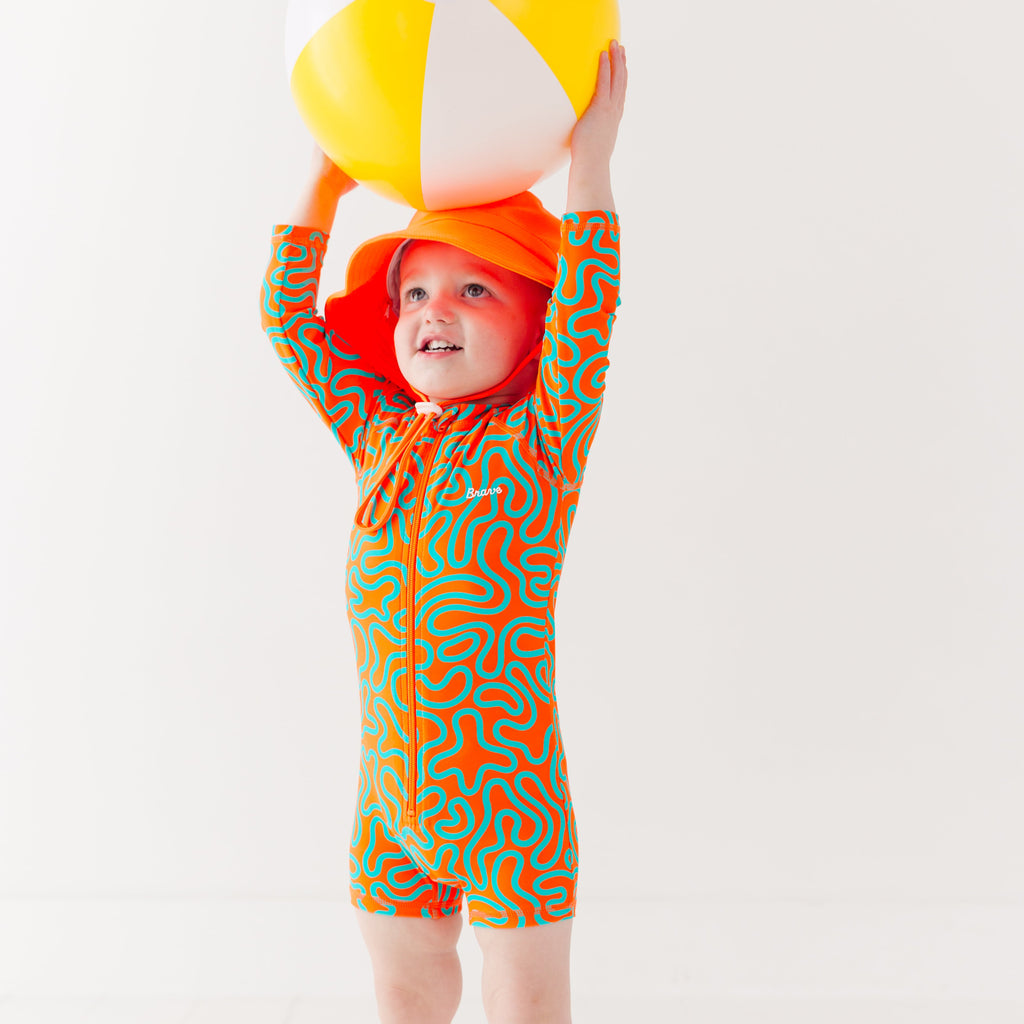 Child wearing a colorful swimsuit with a squiggly pattern and an orange bucket hat on a white background, holding a white and yellow-striped beach ball