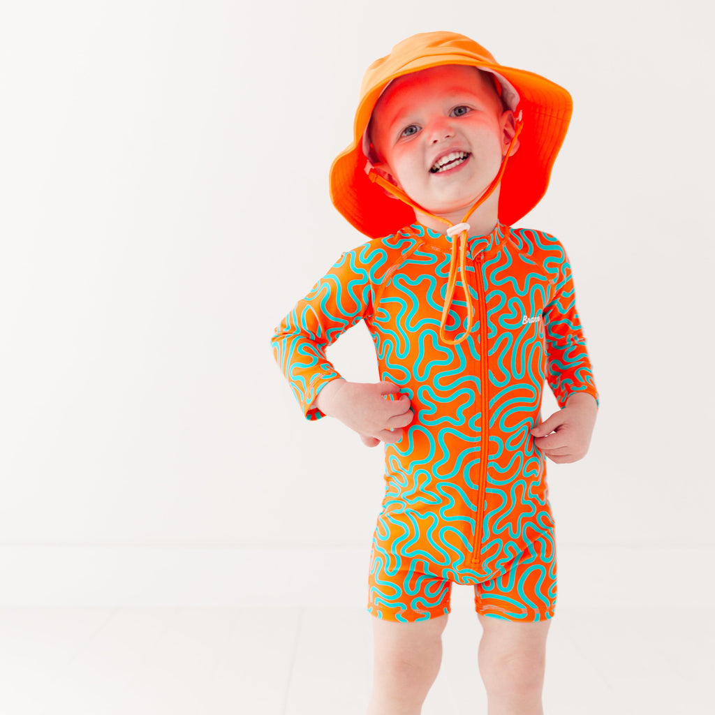 Child wearing a colorful swimsuit with a squiggly pattern and an orange bucket hat on a white background