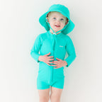 Child wearing a turquoise boy swimsuit and matching bucket hat on a white background