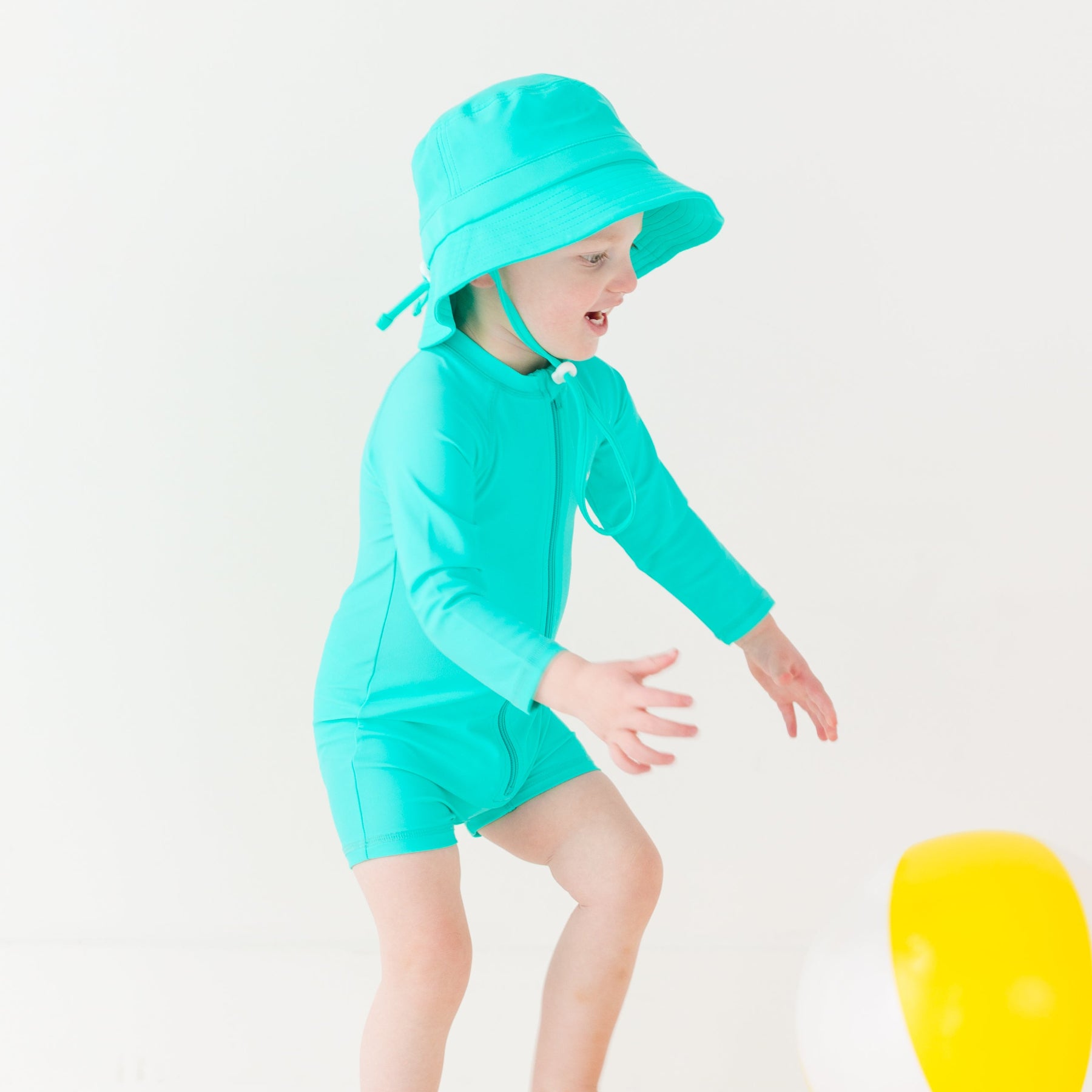 Child wearing a turquoise boy swimsuit and matching bucket hat playing with a yellow beach ball on a white background