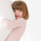 Child in a pink striped long sleeve pajama hugging a white pillow against a white background