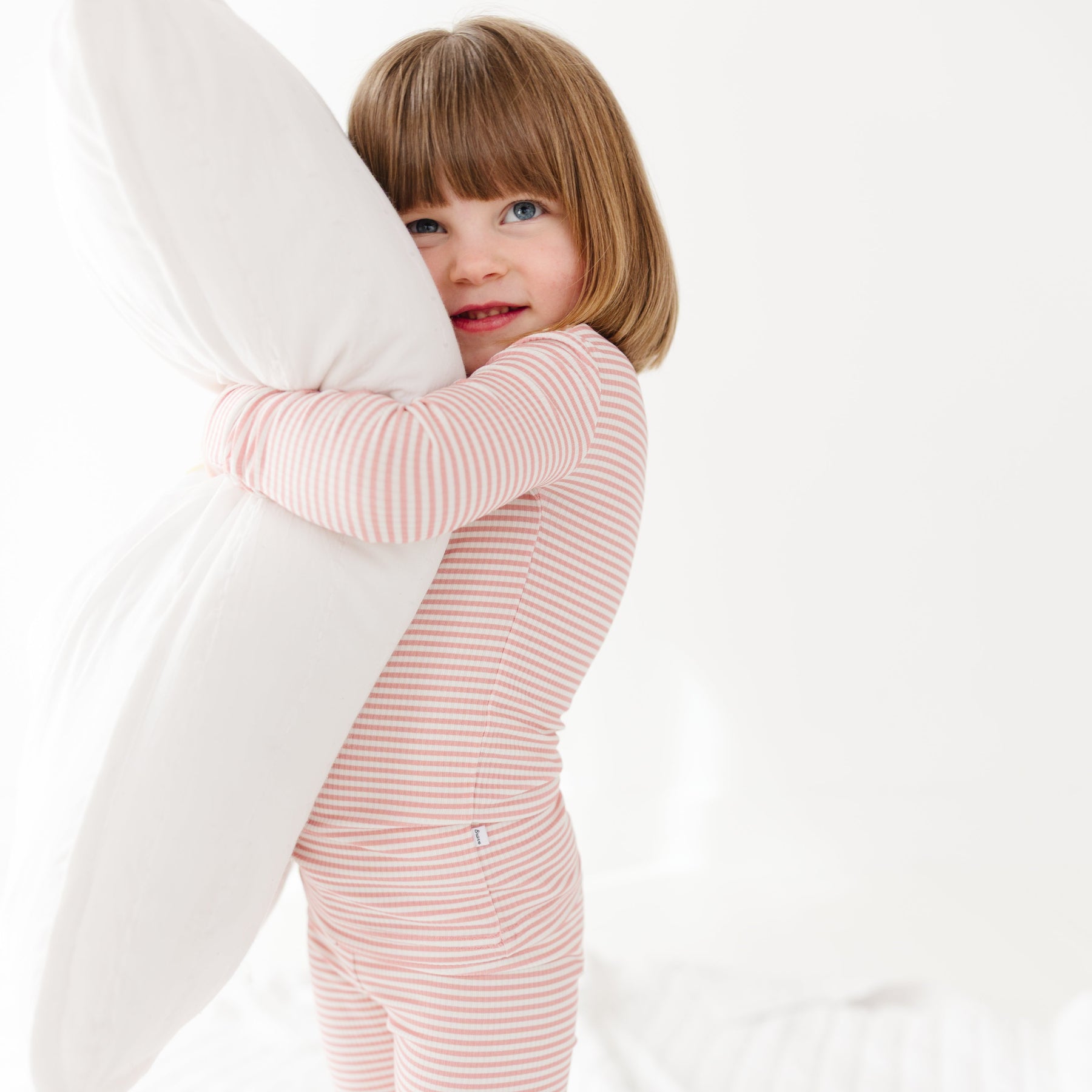 Child in pink striped pajamas holding a white pillow against a white background