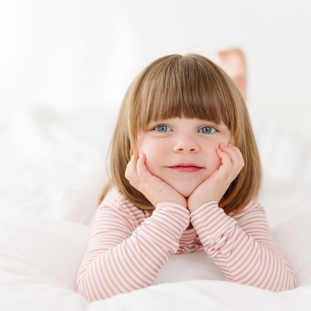 Child wearing a pink striped long sleeve pajama set with her hands on her chin on a white background