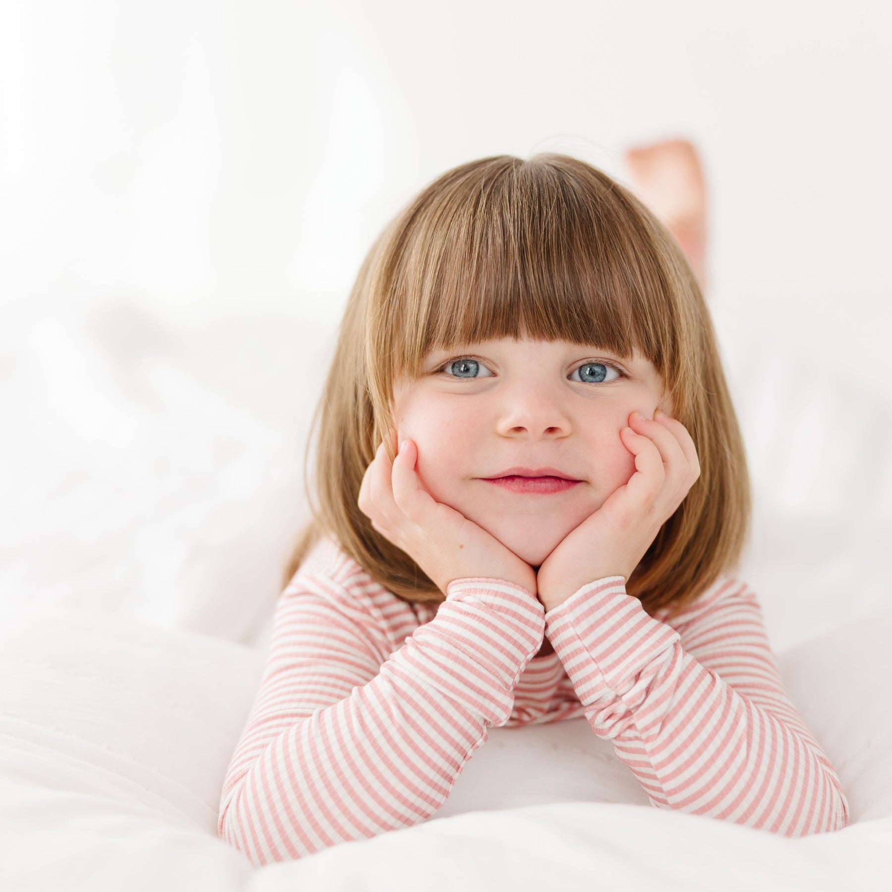 Child wearing a pink striped long sleeve pajama set with her hands on her chin on a white background