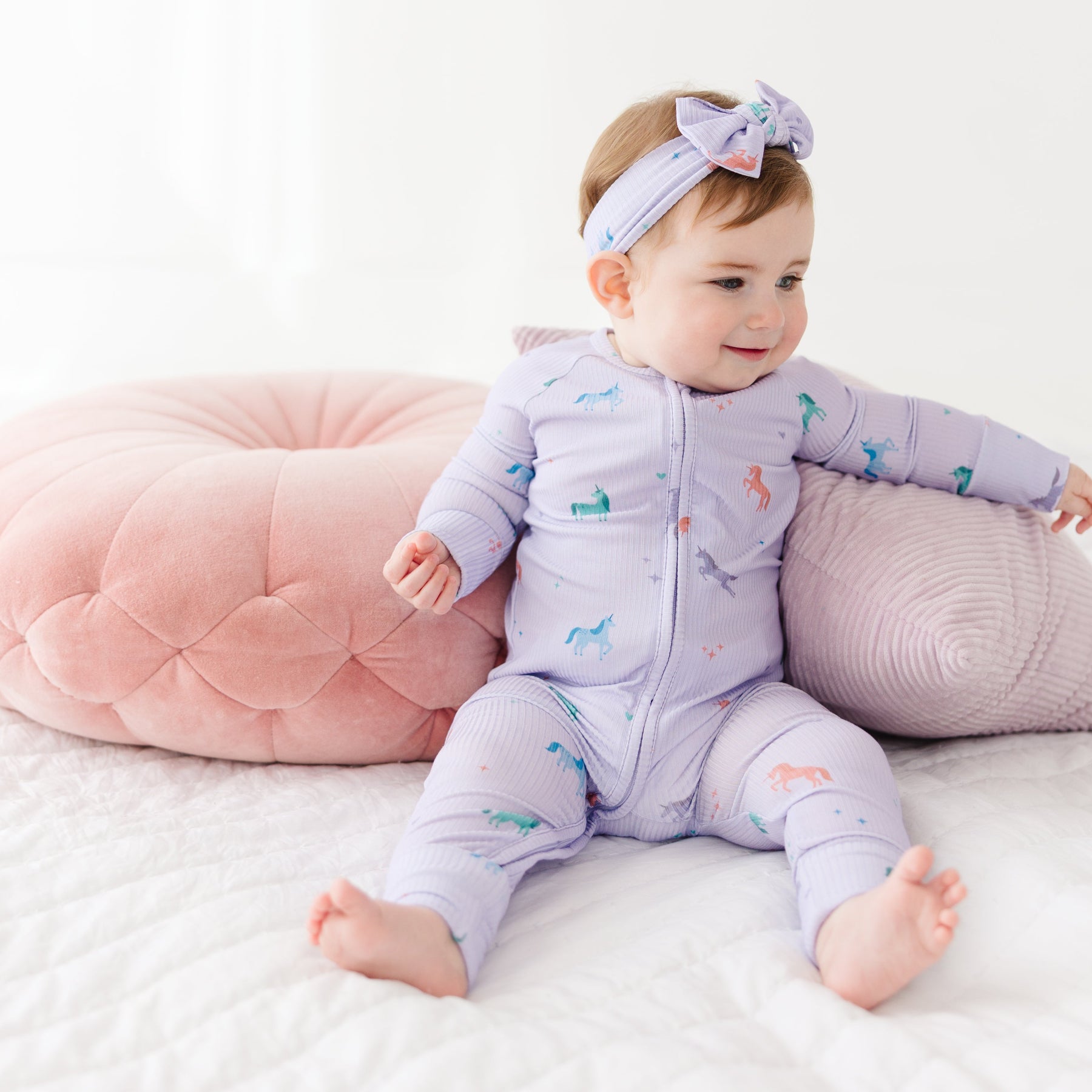 Baby in a purple outfit with animal prints sitting on a white surface with pink and gray cushions.