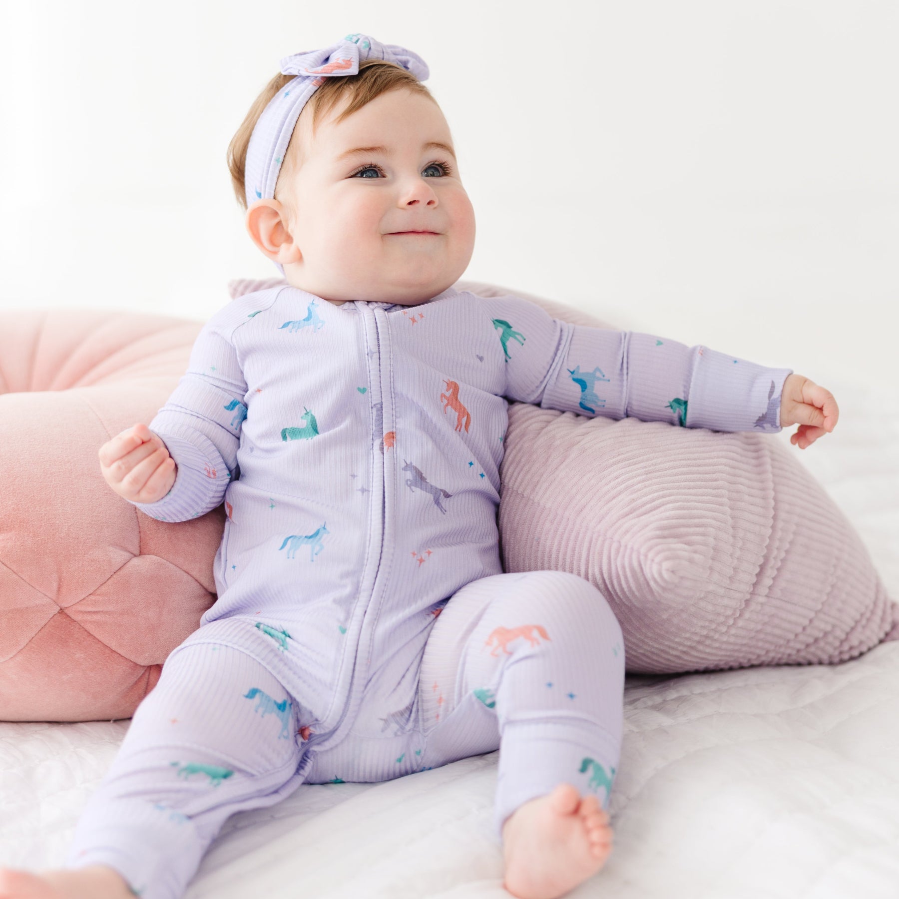 Baby in a purple onesie with animal prints sitting on a white surface with pink pillows.